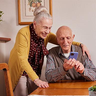 Image Description: an older man sitting at a table while an older woman places her arm around his back.