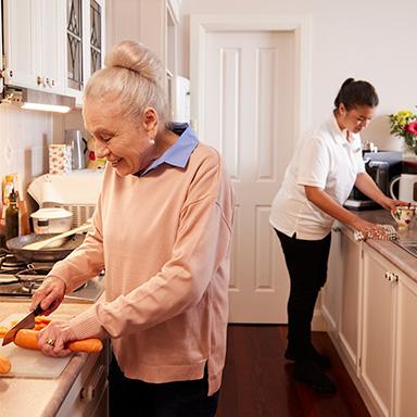 Image Description: aged care worker in kitchen with while older woman chops carrots